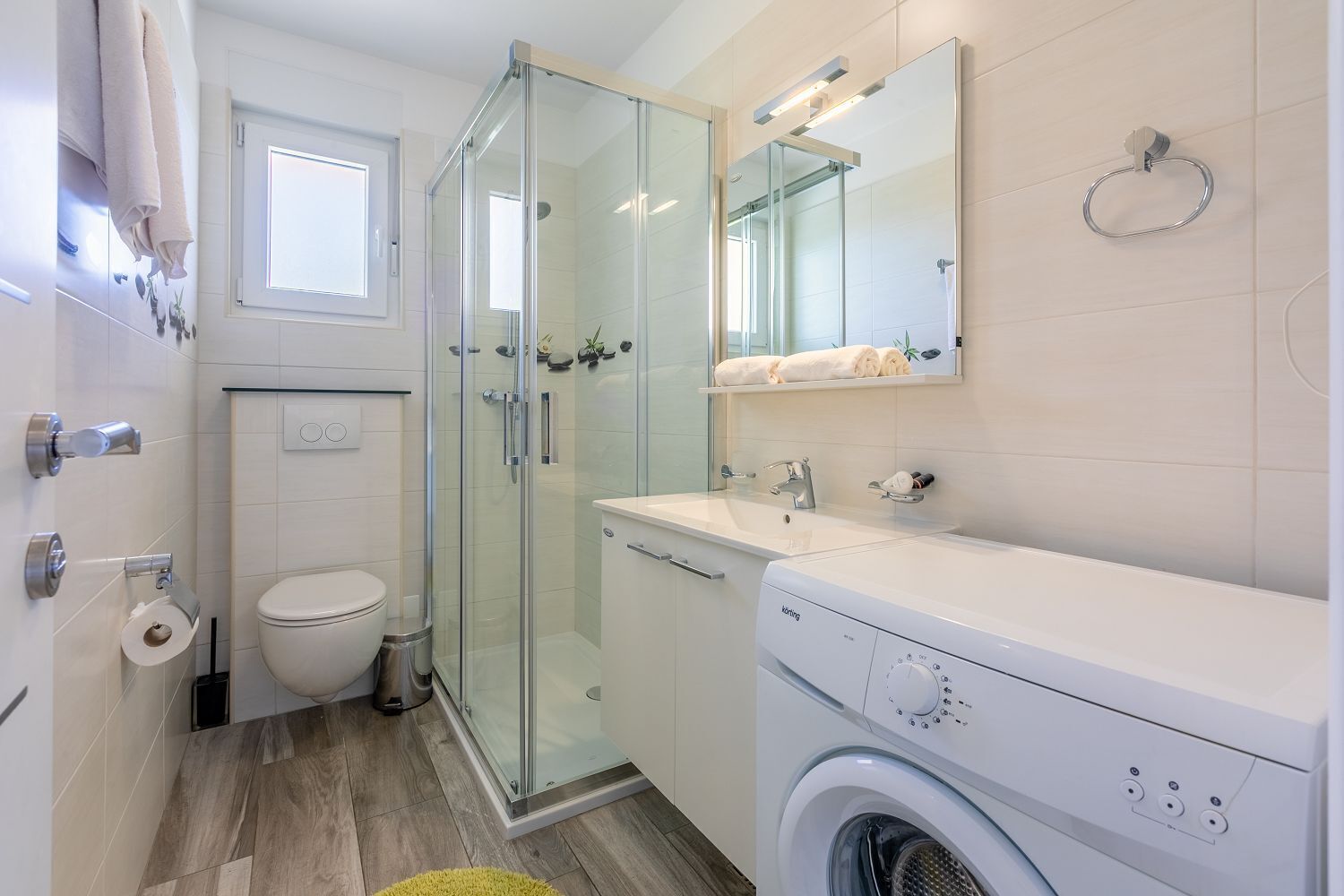 A modern bathroom with a glass-enclosed shower, a toilet, a washing machine, and a sink with a mirror above it.