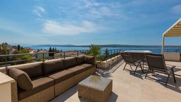 A terrace with a sofa, chairs, and a table overlooking a scenic view of the sea and mountains.