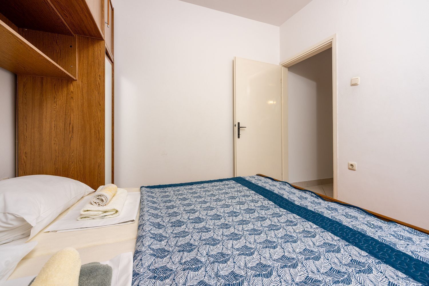 A small, simple room with a bed, a wooden wardrobe, and a door leading to another room.