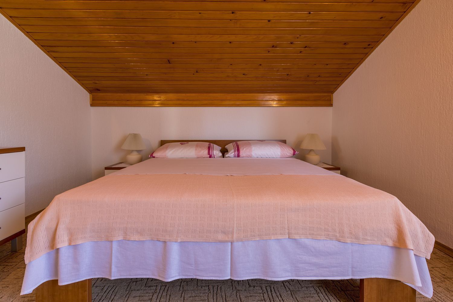 A bedroom with a wooden ceiling, a bed with a pink and white bedspread, two bedside lamps, and a nightstand with a lamp.