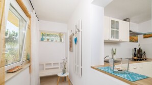 Eine schmale, helle Küche mit weißen Schränken, einem kleinen Esstisch und einem Fenster mit Blick auf Grünflächen.