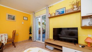 Een woonkamer met een televisie, een eethoek met een tafel en stoel, en een schuifpui naar een buitenruimte.