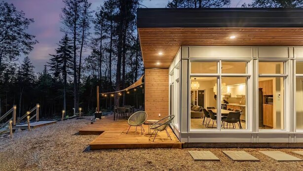 Una cabaña moderna con fachada de madera, amplios ventanales y una terraza con mobiliario exterior.
