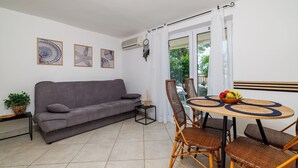 Ein Wohnzimmer mit grauem Sofa, einem kleinen Tisch mit Obstschale, Korbstühlen und einem Fenster mit Vorhängen.
