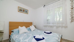 Ein ordentlich gemachtes Bett mit einem Bettbezug mit Blattmuster, ein Nachttisch mit Lampe und ein Fenster mit durchscheinenden Vorhängen.