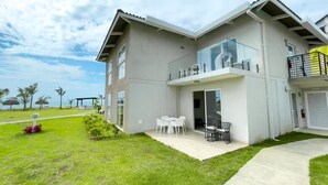 Una casa moderna de dos pisos con balcón, un patio con mobiliario exterior y un césped bien cuidado.