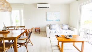 Un salón luminoso con una mesa de comedor de madera, sillas de mimbre, un sofá blanco con cojines estampados y un ventanal con vistas al exterior.