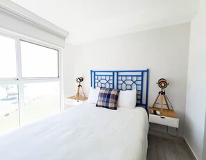 A hotel room with a large bed, a blue headboard, two bedside tables with lamps, and a window providing a view. - Oceanfront Serenity -V5-3E- 2 bed, 2 bath (Punta Chame)