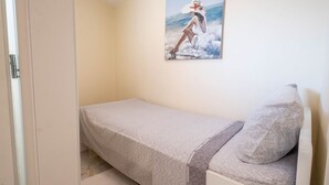 Ein kleines Zimmer mit einem Einzelbett, einer schlichten Bettdecke und einem Kopfkissen. Ein Bild von einer Frau am Meer hängt an der Wand.