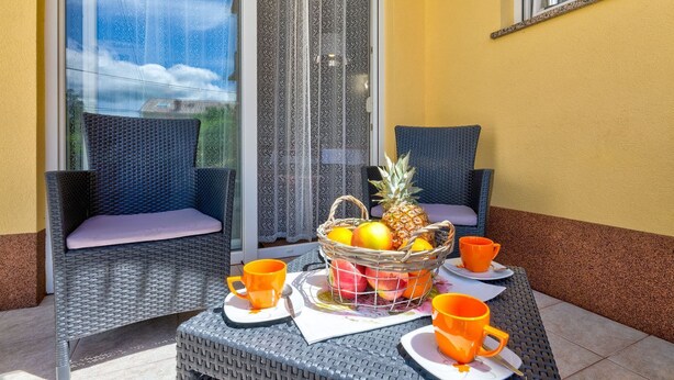 Eine Terrasse mit Korbstühlen, einem Tisch mit orangefarbenen Tassen und einer Obstschale, sowie ein Blick durch eine Glastür.