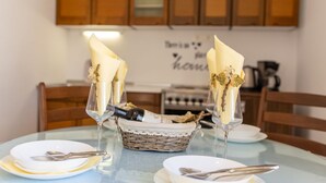 Une table à manger est dressée avec deux verres à vin, chacun accompagné d’une serviette pliée et d’une branche décorative, le tout posé sur des assiettes blanches avec des couverts. Un panier en osier contenant une nappe en dentelle est également posé sur la table.