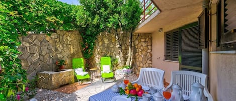 Un patio al aire libre con paredes de piedra, sillas verdes, una mesa dispuesta para comer y diversas plantas y flores.