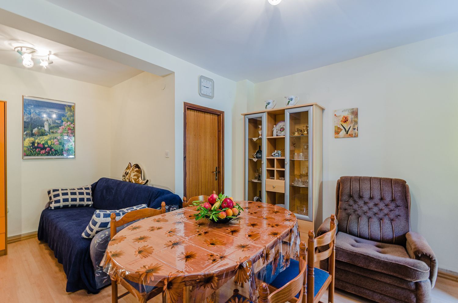 Una sala de estar acogedora con una mesa redonda de madera, un sofá, un sillón con cojines y un aparador con puertas de cristal.
