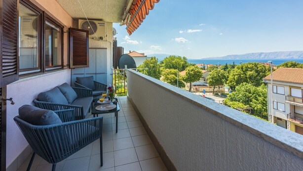 Un balcón con sofá, mesa y sillas, con vista panorámica a un pueblo costero y montañas.