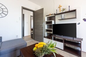 A modern living room with a grey sofa, a wooden coffee table with fruits, a TV on a wooden stand, and a door leading to another room. - Bright apartment just 100 metres from the beach with sea view, parking space, BBQ (Novi Vinodolski)
