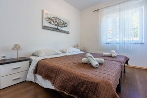 A hotel room with two beds, a bedside table with a lamp, a painting on the wall, and a window with sheer curtains. - Bright apartment just 100 metres from the beach with sea view, parking space, BBQ (Novi Vinodolski)