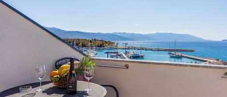 A balcony with a table set for two, overlooking a marina with boats and a mountainous backdrop.