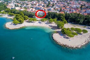 An aerial view of a coastal area with a small island, clear turquoise waters, and a densely built urban area in the background. - Fantastic apartment right on the beach promenade with lots of palm trees (Crikvenica)