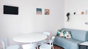 Ett modernt vardagsrum med ett runt, vitt bord, fyra vita stolar, en blå soffa med blommiga kuddar och en platt-TV på väggen.