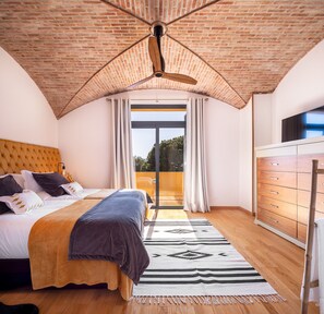 Une chambre à coucher avec un lit, une commode en bois, un ventilateur de plafond et une vue sur l’extérieur, visible à travers une porte vitrée.