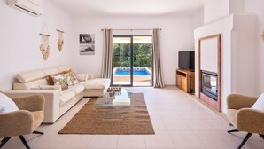 Ein modernes Wohnzimmer mit einer Couch, einem Couchtisch, Stühlen, einem Fernseher und Blick auf einen Swimmingpool durch Glastüren.