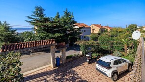 Zona residencial con un coche blanco estacionado frente a una casa, un espacio de estar al aire libre cubierto y vista al mar al fondo.