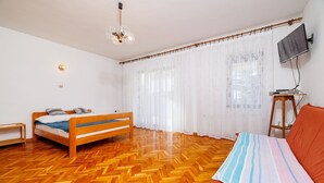 Un dormitorio con cama y suelo de madera, un televisor en la pared y una ventana con cortinas ligeras.