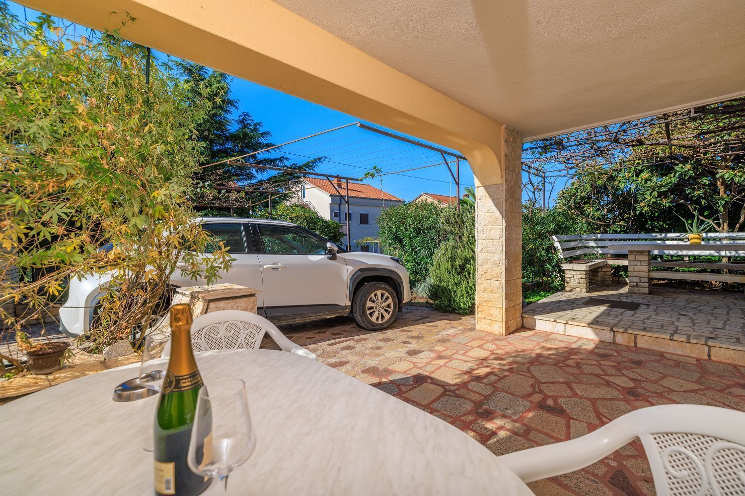 Un patio techado al aire libre con una mesa lista para comer, un coche aparcado y un banco.