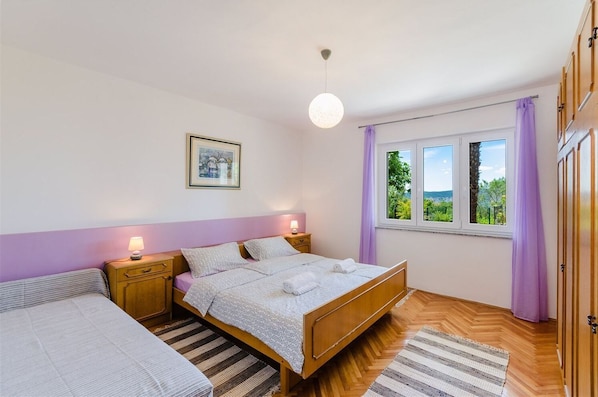 A bedroom with a bed, a nightstand, a lamp, a window with curtains, and a view of greenery outside. - Mediterranean apartment with sea view, parking space, cot, garden (Crikvenica)