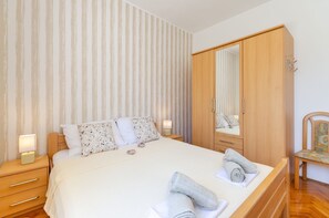 Uma cama bem arrumada com edredom branco, um guarda-roupa de madeira e uma cadeira com almofada estampada.