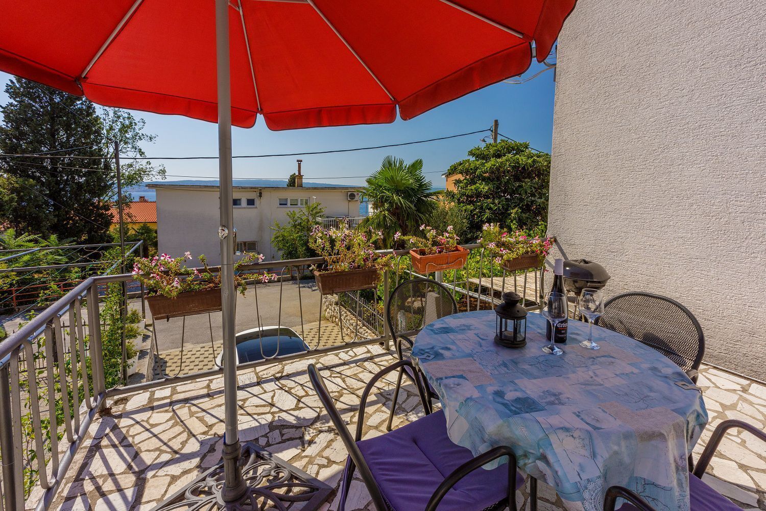 En balkon med et bord, stole og en rød parasol. Der er krukker med planter og udsigt til bygninger i baggrunden.