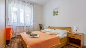 Een slaapkamer met een bed, nachtkastje, lamp, raam met dunne gordijnen en een schilderij aan de muur.