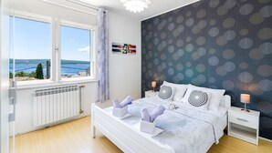 A bedroom with a large window, a bed with patterned pillows, a nightstand, and a wall with a circular pattern. - Bright apartment just 200 metres from the sandy beach with sea views (Crikvenica)