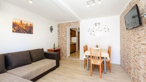 Un salon moderne avec un canapé, une table à manger et des chaises. On aperçoit au fond une cuisine, un mur en briques et un téléviseur fixé au mur.