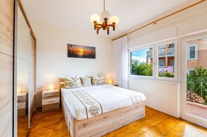 A bedroom with a wooden bed, a nightstand, a bedside table, a mirror, a chandelier, a window with a view of a balcony, and a painting on the wall. - Vintage-style apartment just 450 metres from the beach with sea views – dogs welcome (Crikvenica)