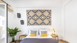Quarto moderno com uma cama grande, duas mesinhas de cabeceira, uma obra de arte na parede, um vaso de planta e uma janela.