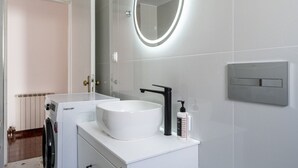 Uma casa de banho moderna com uma pia branca, um espelho redondo com luz, uma torneira preta e um interruptor de luz fixado na parede.