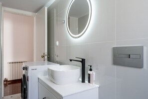 A modern bathroom with a white sink, a round mirror with a light, a black faucet, and a wall-mounted light switch. - Massive 4BR Retro Apartment | 3 min Walk to Beach (Kos)