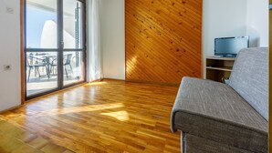 Una stanza con pavimento in legno, un divano grigio, un televisore su un mobile in legno e una porta scorrevole in vetro che dà su una zona esterna con sedie.