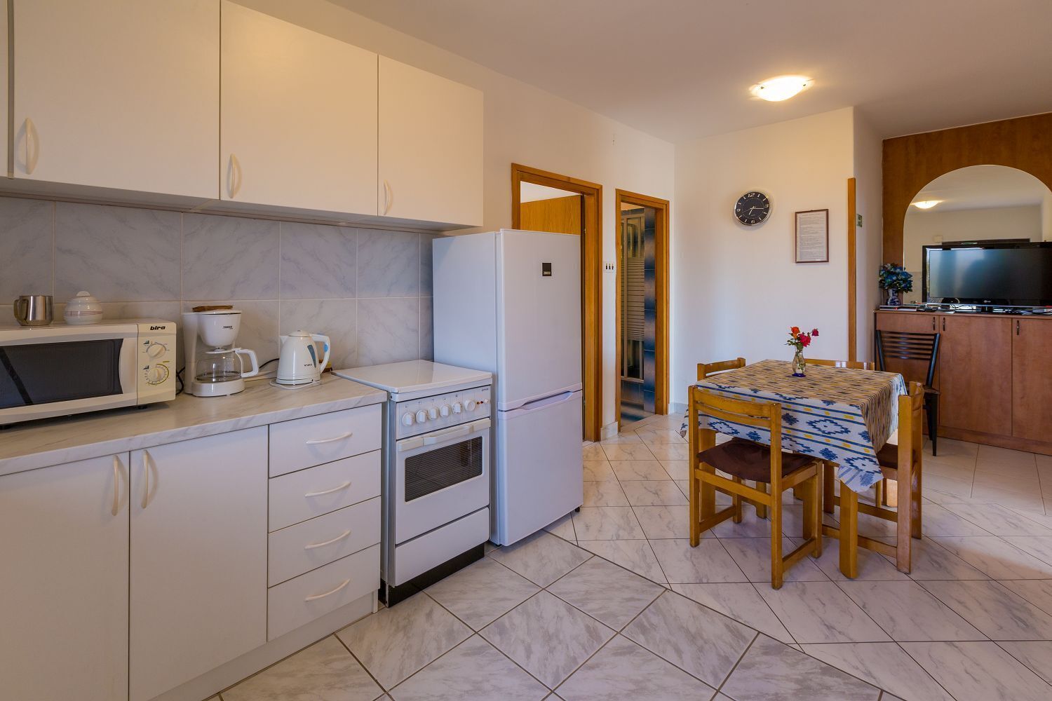 Cocina compacta con microondas, cafetera y horno. Se observa una mesa de comedor con sillas y un televisor al fondo.