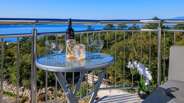 Een ronde tafel met een fles wijn, twee glazen en een vaas met bloemen, met op de achtergrond een helderblauwe lucht en een wateroppervlak.