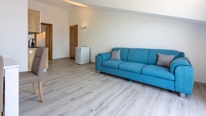 Een moderne woonkamer met een blauwe bank, een houten stoel, een kleine witte koelkast, en een keuken die zichtbaar is door een open deur.