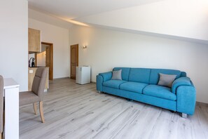 Ein modernes Wohnzimmer mit einem blauen Sofa, einem Holzstuhl, einem kleinen, weißen Kühlschrank und einem durchgehenden Türrahmen erkennbaren Küchenbereich.