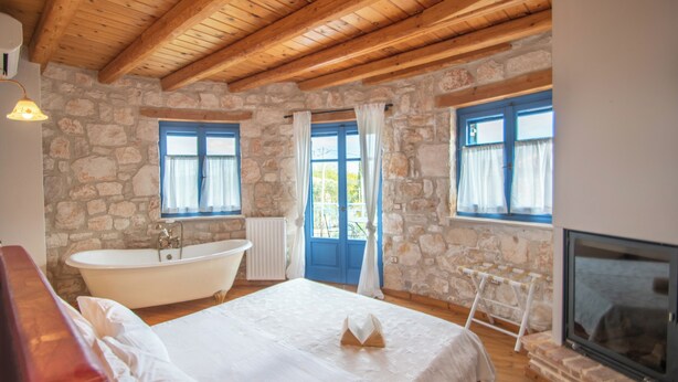 A bedroom with a stone wall, wooden ceiling beams, a bathtub, and a fireplace. - Valory’s Stone Tower (Ano Vasilikos)