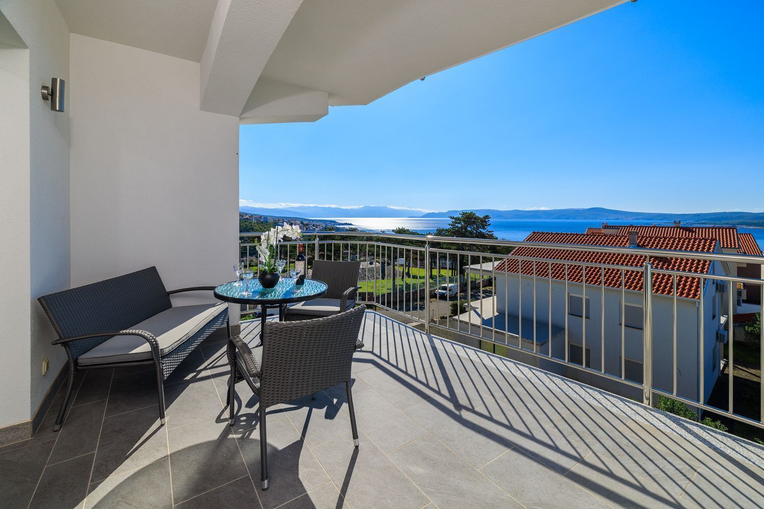 Un balcone con tavolo e sedie, affaccia su una vista costiera con edifici e cielo sereno.