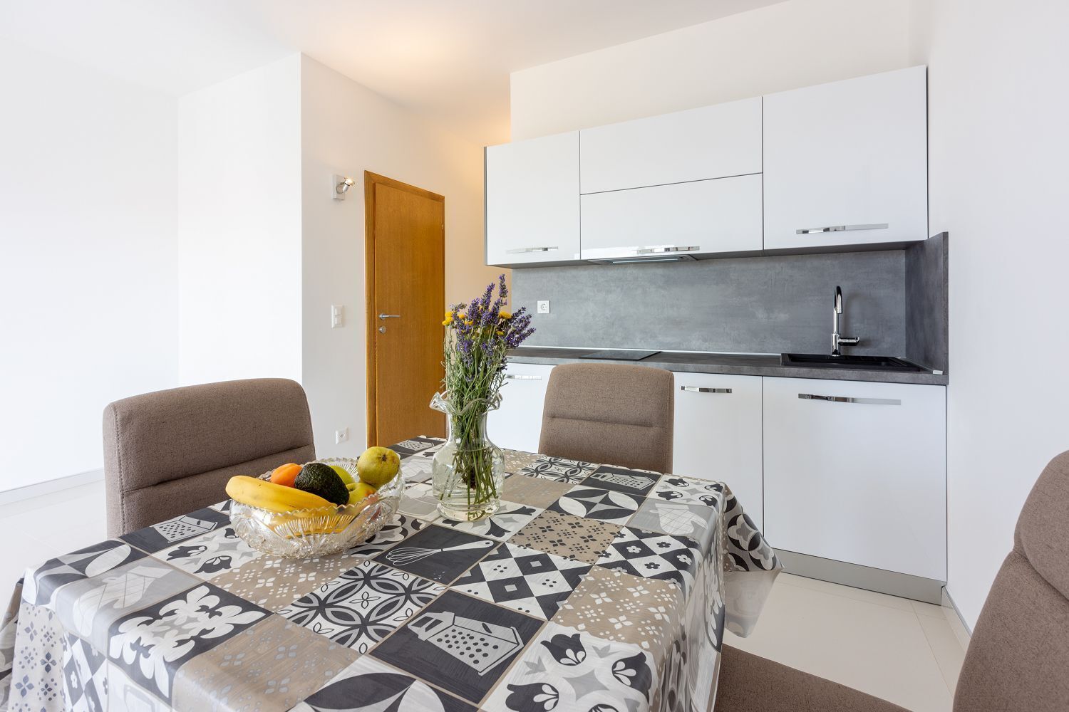 A modern kitchen with white cabinets, a built-in oven, and a sink. There is a dining table with a patterned tablecloth and a vase with flowers.