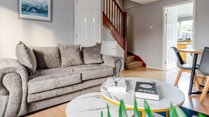 Una sala de estar con un sofá gris, una mesa de centro con revistas, una escalera y una puerta que da a otra habitación.