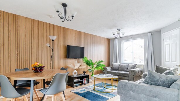 Una sala moderna con pared de paneles de madera, un sofá gris, una mesa de comedor con sillas, una mesa de centro y un televisor montado en la pared.
