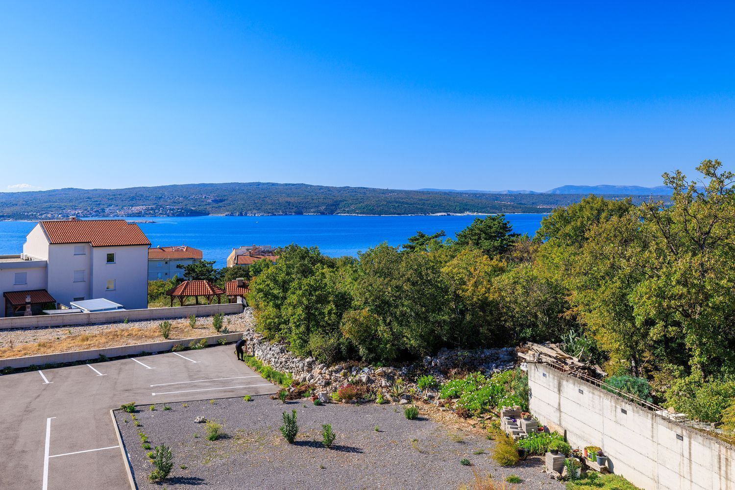 A coastal residential area with a parking lot, a small building with a red roof, and a view of the sea and distant hills.