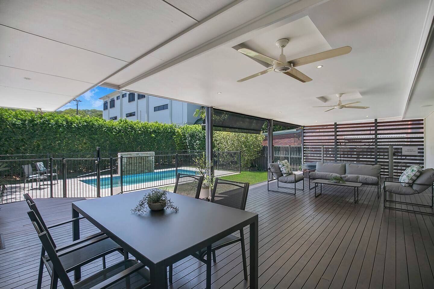 A spacious, covered deck designed for easy alfresco living, with a comfortable lounge and dining setting beneath ceiling fans, overlooking the private backyard and pool for relaxed meals, shaded downtime, and long afternoons outdoors.